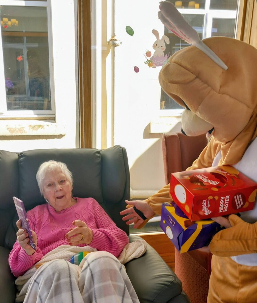 An elderly woman sits at Lear House holding a chocolate bar as someone in an Easter Bunny costume hands her Easter eggs indoors during an Egg-stra Special Easter Celebration.