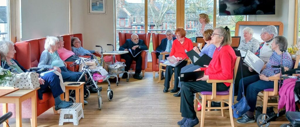 A group of elderly people sit in a common room, some holding songbooks, taking part in a social or musical activity together.
