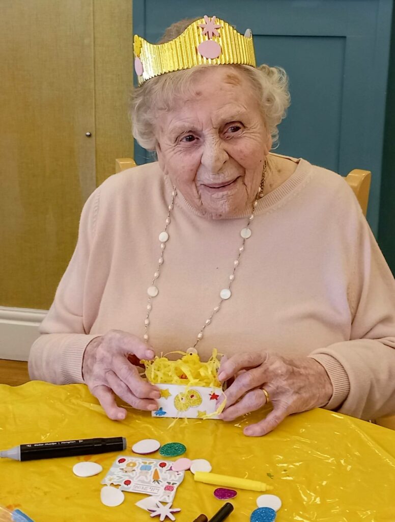Elderly woman wearing a yellow paper crown sits at a table with arts and crafts materials, holding a decorated box and smiling.