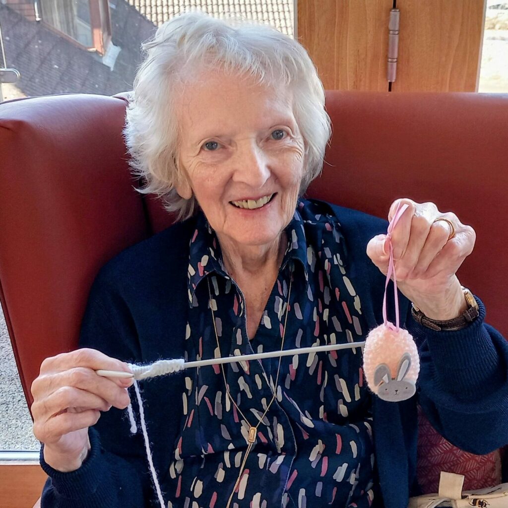 An elderly woman sits in a chair, stitching with white wool and holding up a special pink bunny-shaped ornament—just right for Easter.