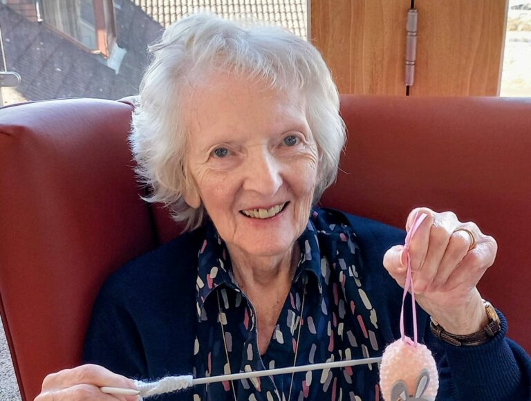 An elderly woman sits in a chair, stitching with white wool and holding up a special pink bunny-shaped ornament—just right for Easter.