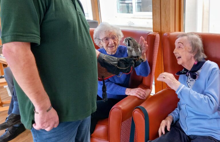 A man holds a small owl by its talons on his gloved hand, while two elderly women seated in chairs enjoy this unique visit with the bird inside a bright room.