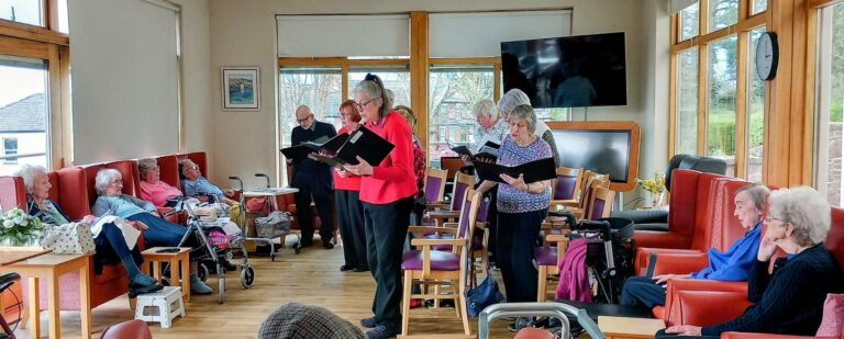 A small choir performs for a group of elderly residents seated in a bright, communal room with large windows.
