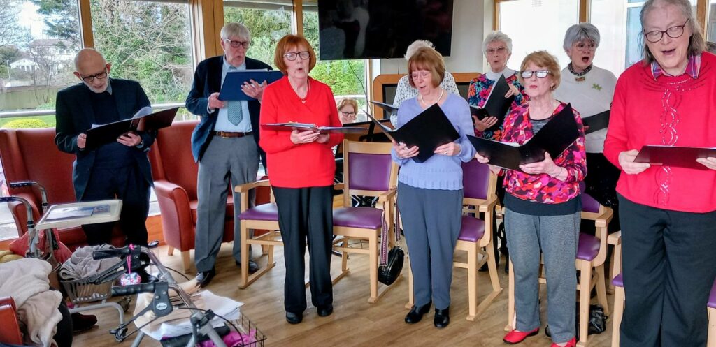 A group of older adults stand in a room holding sheet music, singing together. Some are wearing spectacles. Several empty chairs and a walking frame are visible in the foreground.