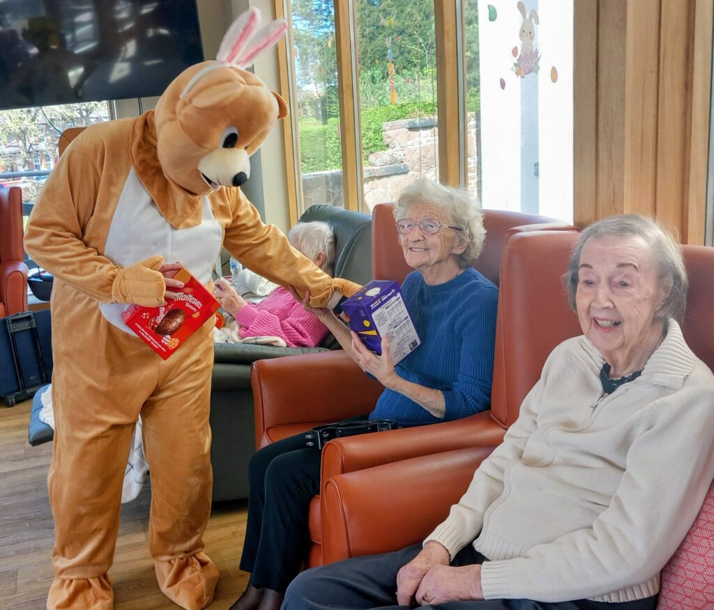 During the Egg-stra Special Easter Celebration at Lear House, a person in an Easter Bunny costume hands out chocolate eggs to two smiling elderly women seated in a lounge area.