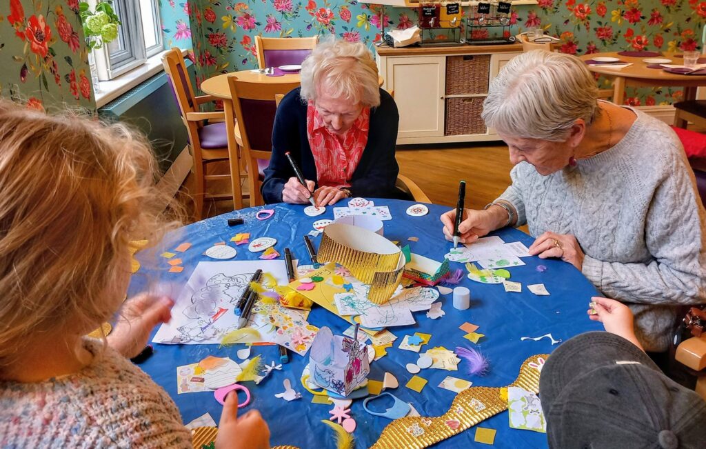 Two elderly women and two children sit around a table covered with craft materials, drawing and creating art together in a brightly decorated room.
