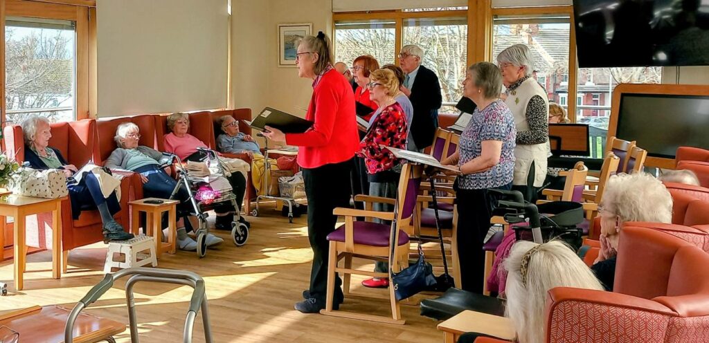 A small group of older adults stands singing in a brightly lit common room, while other pensioners sit and listen from chairs and settees.