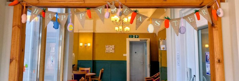 A doorway adorned with a special Happy Easter banner featuring fabric carrots and eggs, detailed with delicate stitching, opens into a room containing tables, chairs, and a ‘Home Sweet Home’ sign on the wall.