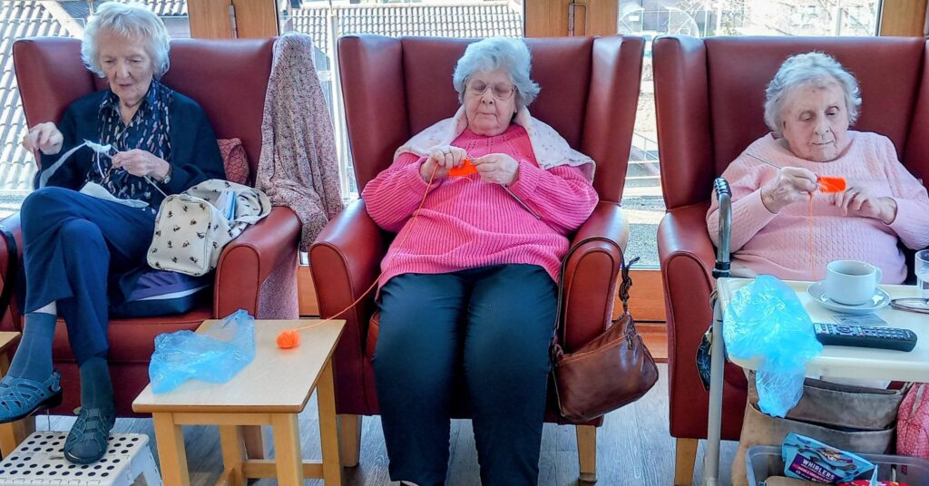 Three elderly women sit in armchairs, each knitting with orange wool. A small table holding a bag, a cup, and a remote sits beside them in a sunlit room, creating a particularly inviting and cosy atmosphere.