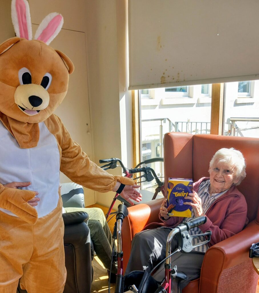 During an Easter celebration at Lear House, someone in a bunny costume stands beside an elderly lady who smiles whilst holding a large box of Cadbury Twirl chocolate for an egg-stra special moment.