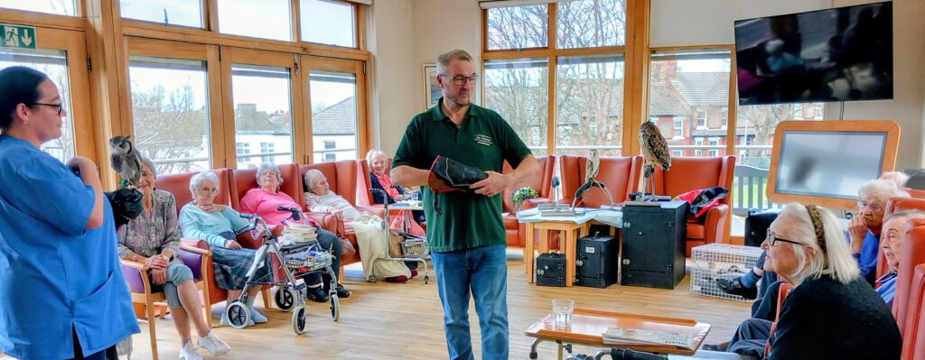 A man presents a bird demonstration to elderly residents in a care home lounge during a special visit, as a woman holds an owl with sharp talons and several other birds can be seen nearby.