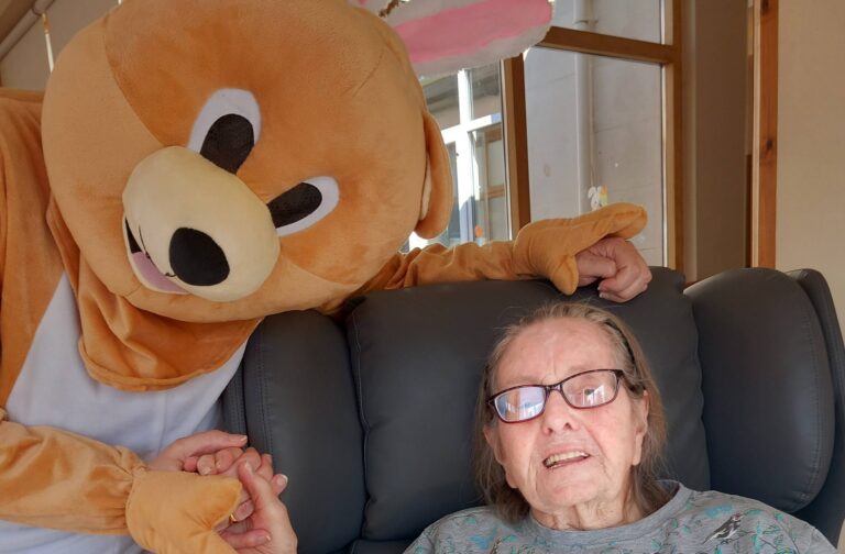 At Lear House, an elderly woman sits in an armchair holding hands with someone in a large bear costume, sharing an Egg-stra Special moment during the Easter Celebration in a sunlit room.
