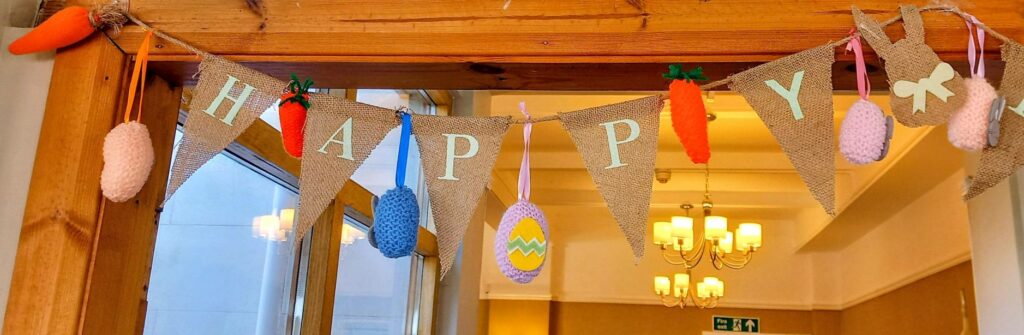 Hessian bunting with letters spelling HAPPY, adorned with soft carrots and eggs, displays delightful Easter crafts and hangs in a room featuring wooden beams and chandeliers.