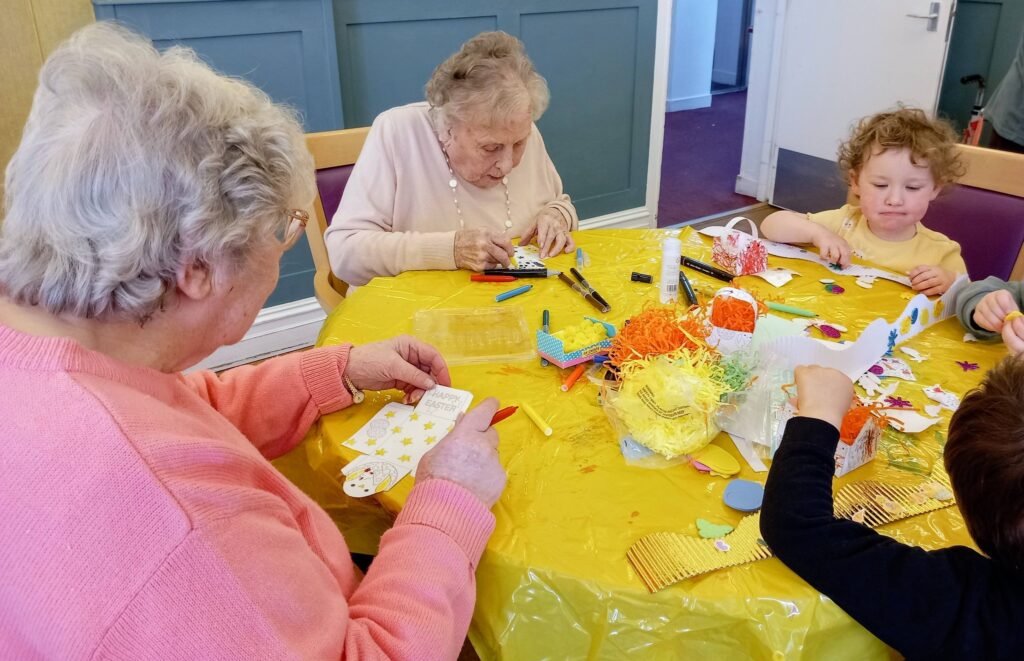 Two elderly women and two children sit at a table draped with a yellow cloth, creating crafts using paper, felt-tip pens, and decorations.
