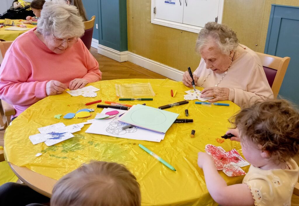 Two elderly women and two young children sit around a table, colouring and decorating paper crafts with felt-tip pens and art materials.