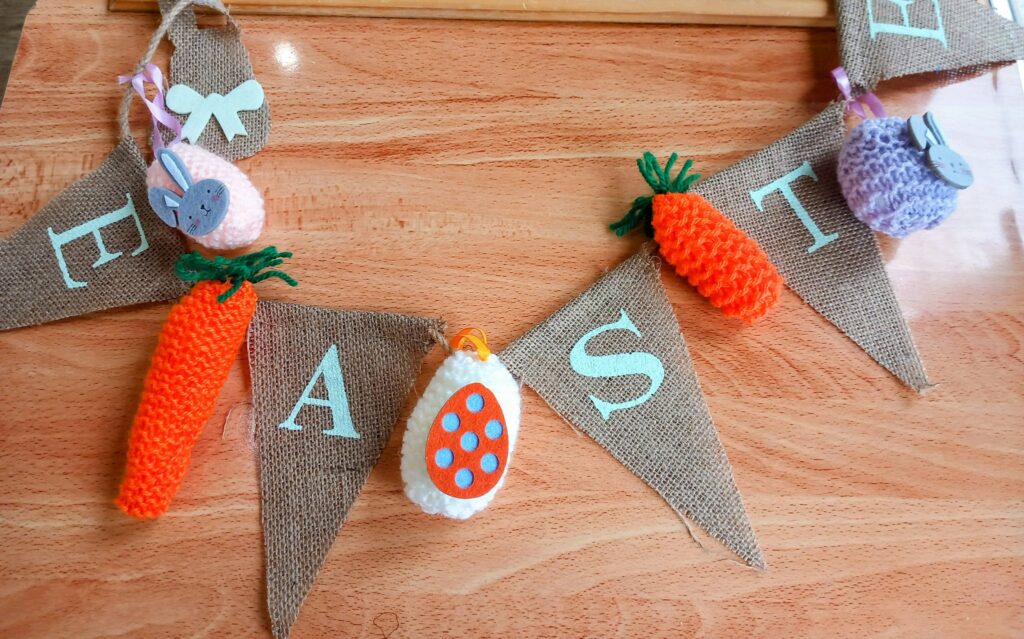 Hessian Easter bunting with white lettering, adorned with intricately knitted carrots, eggs, and animals, set on a wooden surface and finished with delicate stitching.