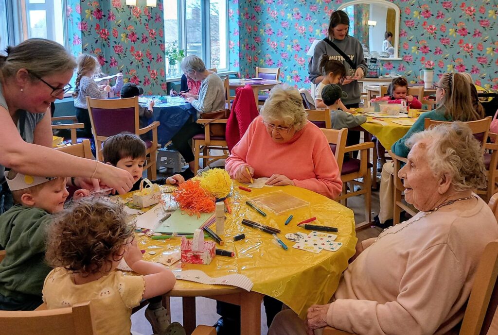 Children and elderly people sit together at tables doing arts and crafts in a brightly coloured room with floral wallpaper.