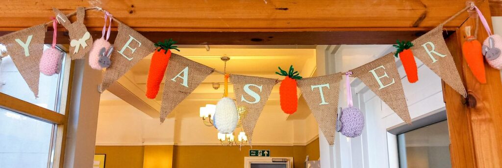 Special bunting spells EASTER in white letters, adorned with knitted eggs, bunnies, and carrots. It features charming stitching and hangs indoors above a doorway.