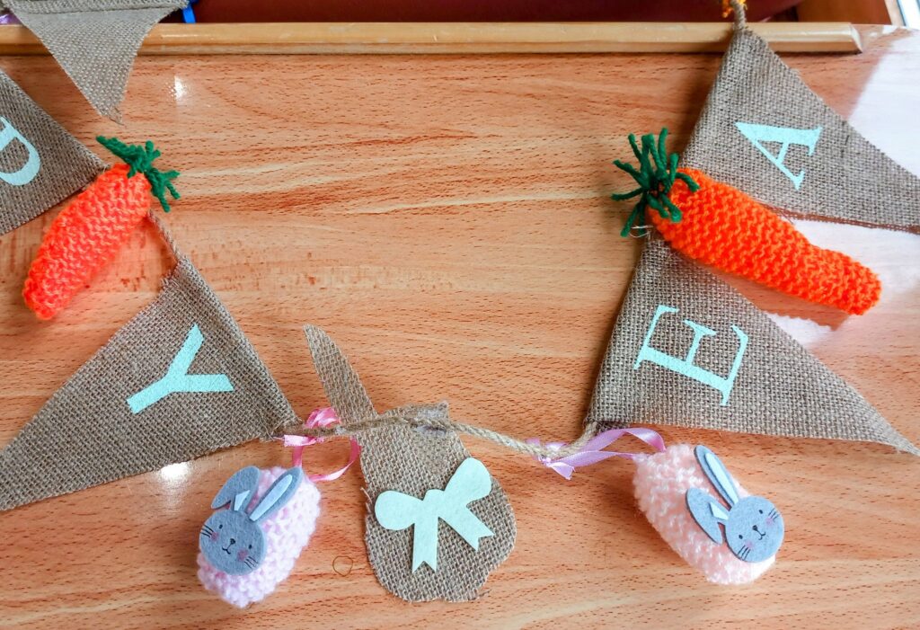 A unique triangular hessian bunting with stitched letters Y, E, and A, knitted carrot decorations, and pink and white bunny ornaments hanging on a wooden surface—ideal for Easter displays.