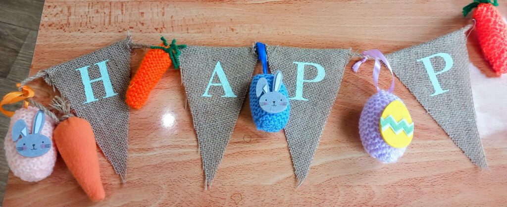 A hessian banner with special stitching spells HAPP, adorned with knitted carrots, rabbits, and an Easter egg, displayed on a wooden surface.