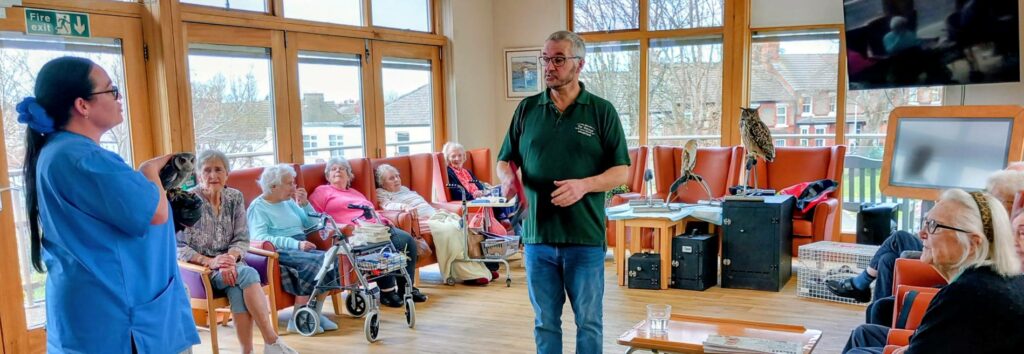 A man addresses a group of elderly people seated in a care home lounge during a special visit, whilst a woman in a blue uniform holds a bird with striking talons.