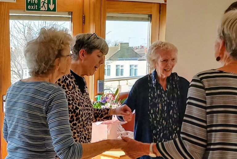 Four older women stand in a sunlit room, holding hands in a circle with gentle smiles. A sense of rhythm connects them, whilst a small table with flowers brightens the background near the window.