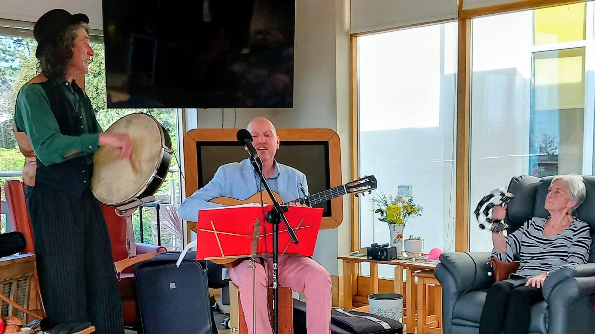 Three people in a well-lit room: one plays a drum with lively rhythm, another sings and plays guitar, both smiling, while the third relaxes in an armchair holding a small dog. Musical gear lies on the floor.
