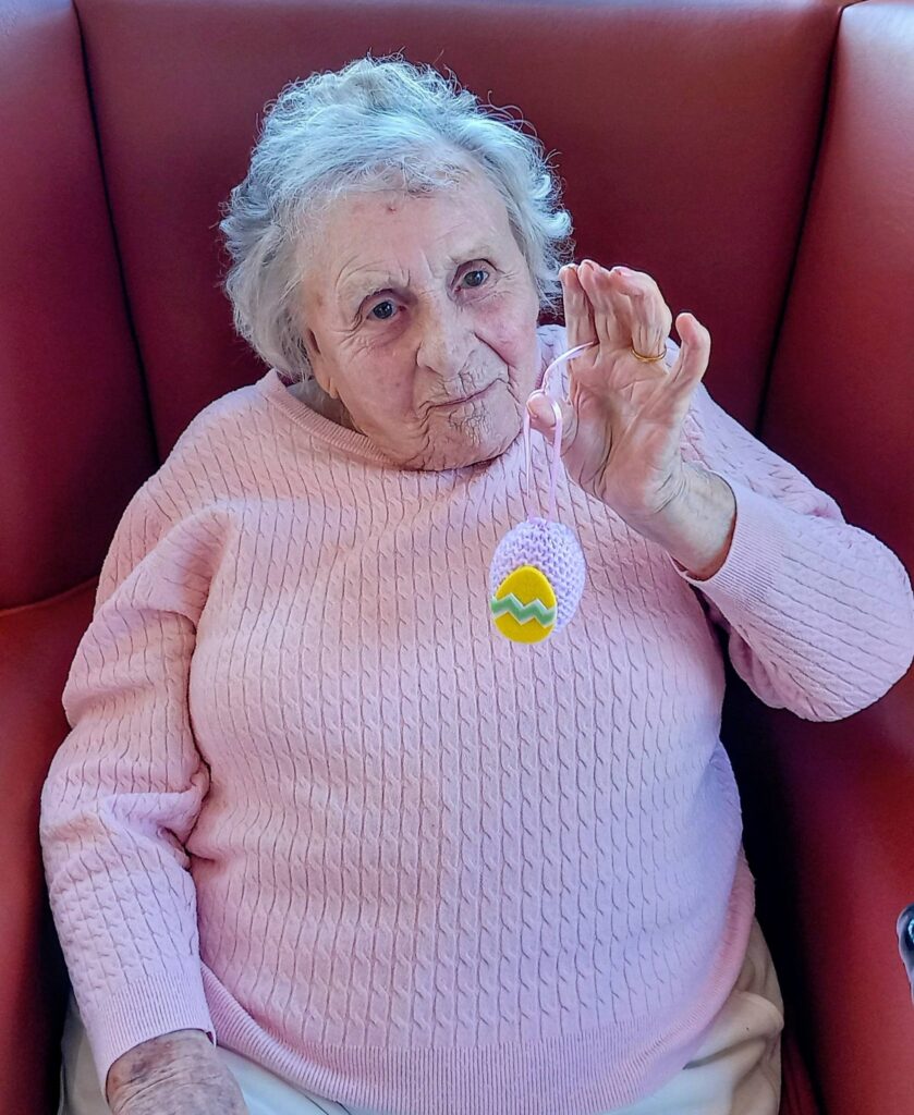 Elderly woman in a pink jumper sits in a red armchair, holding up a special Easter egg decoration featuring a yellow zigzag pattern.