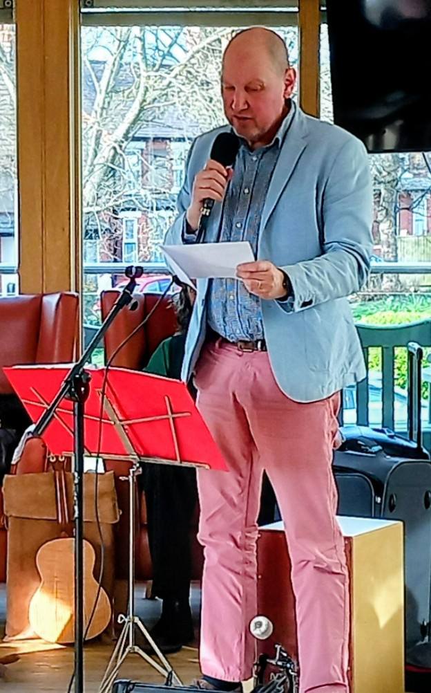 A man in a light blue blazer and pink trousers stands indoors, smiling as he holds a microphone and reads from a sheet of paper, with music stands and instruments nearby.