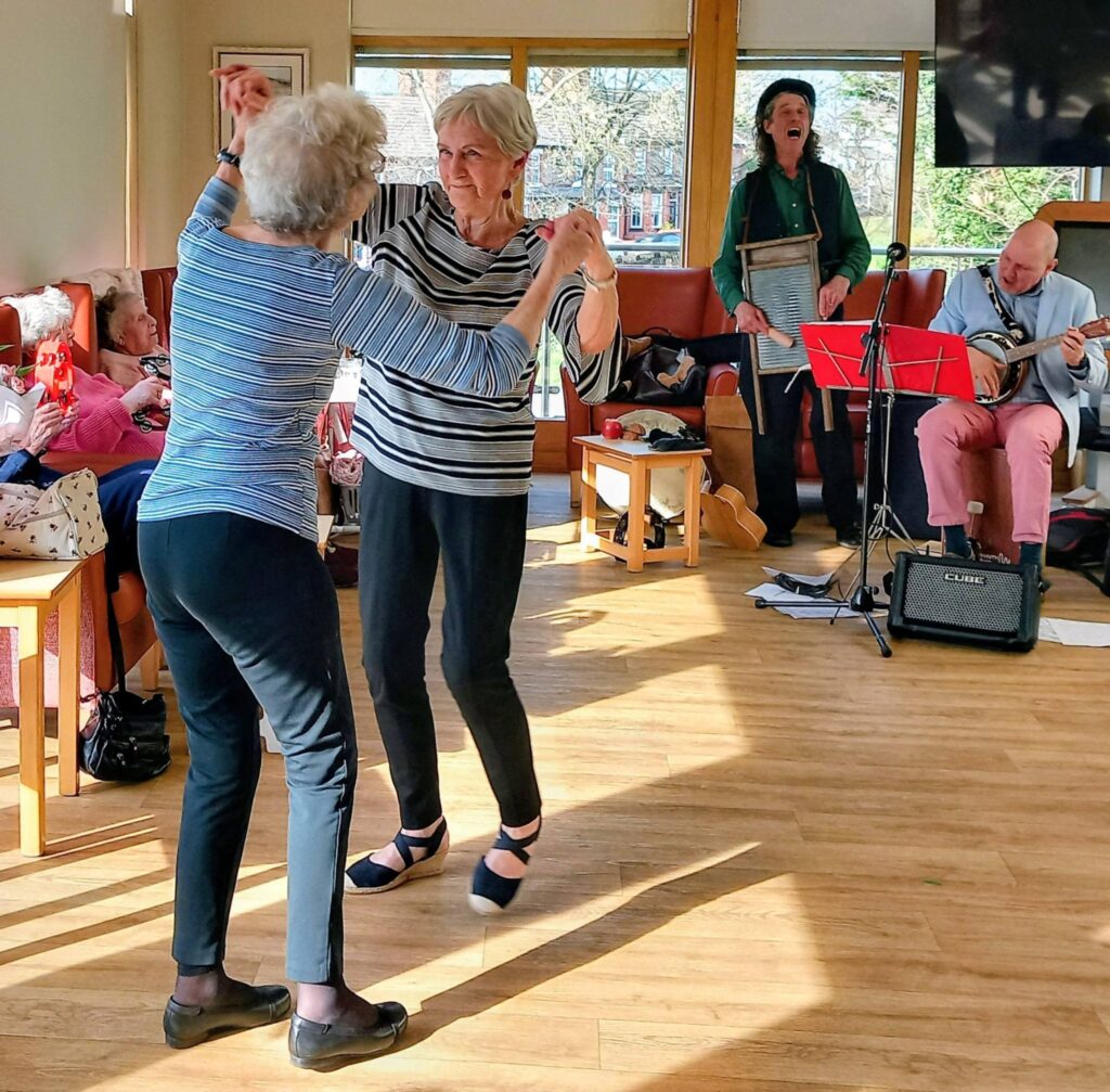 Two older women dance together in a sunlit room, their smiles radiant as they move to the rhythm, whilst a small band with a washboard and guitar plays in the background.