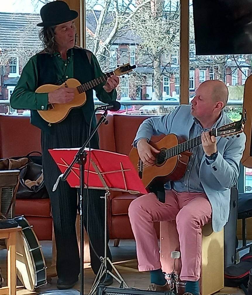 Two musicians perform indoors; one stands playing a ukulele with a smile, whilst the other sits playing a guitar. A music stand and a banjo are visible. Trees and buildings can be seen through the window behind them.