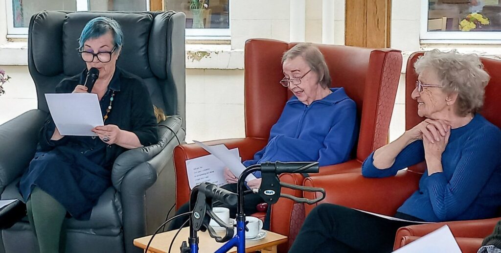 Three older women sit in armchairs, holding papers. One speaks into a microphone whilst the others listen, sharing a thoughtful moment together; a mobility aid is visible in the foreground.