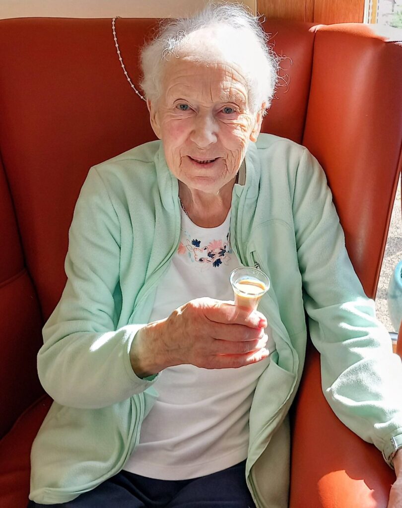 An elderly woman sits in an orange chair, smiling and holding a small glass of creamy drink, enjoying a reflective moment. She wears a light green jacket over a white vest.