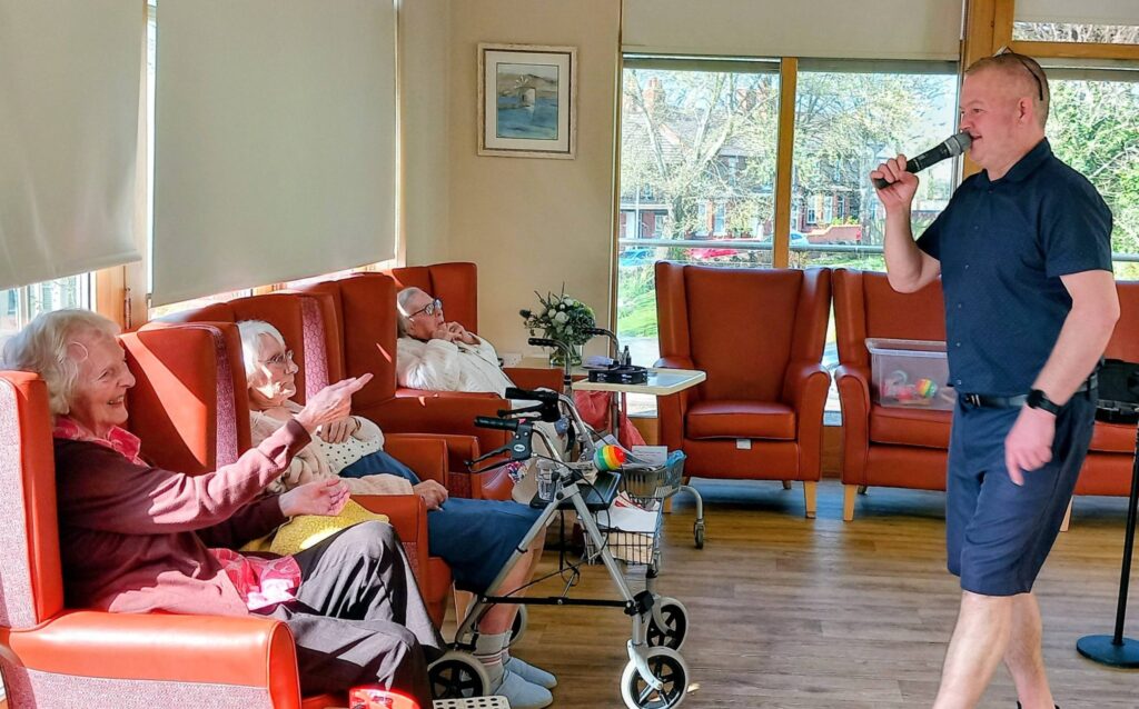 A man in shorts sings into a microphone, creating feel-good fun for a group of elderly people seated in armchairs, enjoying each other's company in a sunlit room.