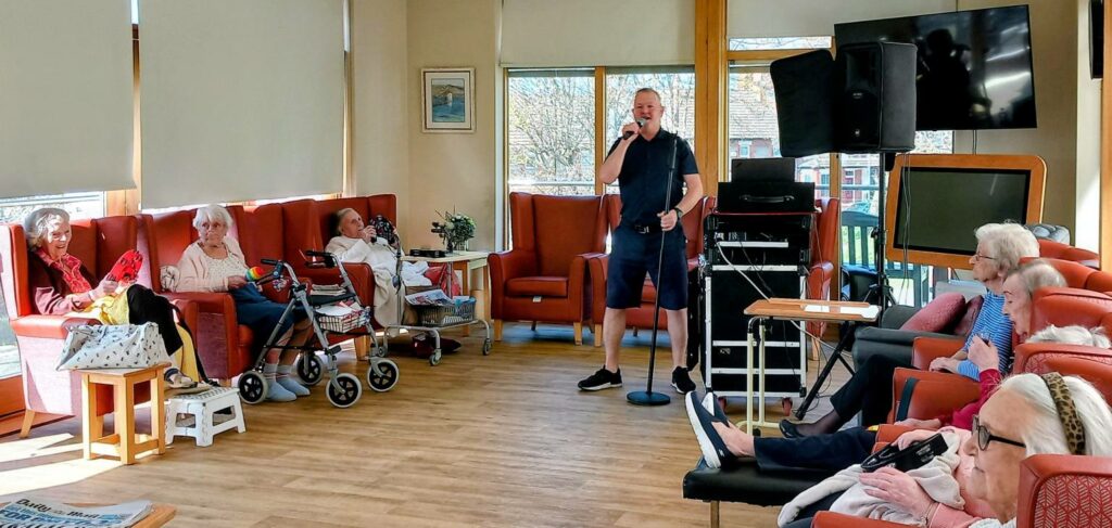 A man sings into a microphone in a care home lounge as several elderly people enjoy the uplifting atmosphere from their armchairs, sharing a meaningful moment together.