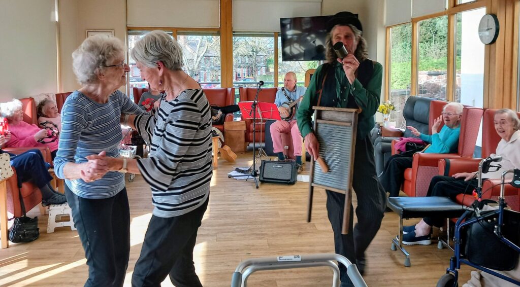 Elderly people in a common room; two women dance as a man in a hat plays the washboard. Smiles are everywhere as residents enjoy the rhythm, with musicians playing gently in the background.