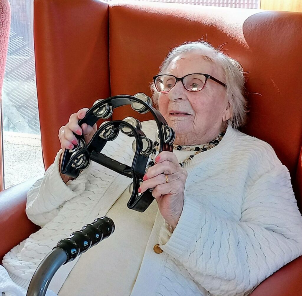 Elderly woman sitting in an armchair, holding a black tambourine and smiling, enjoys some feel-good fun. She is wearing glasses, a white jumper, and has a walking frame in front of her.