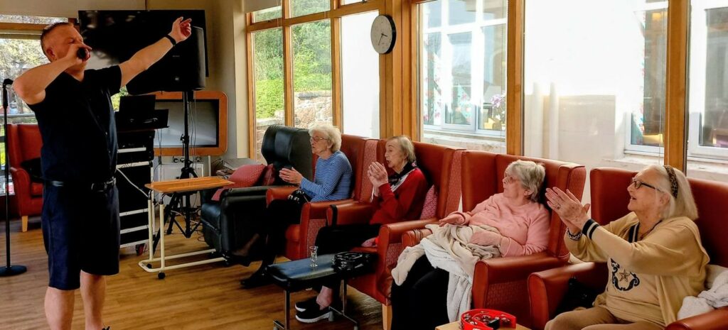 A man sings into a microphone as four older women seated in armchairs clap along, sharing joyful moments in a brightly lit room with large windows.