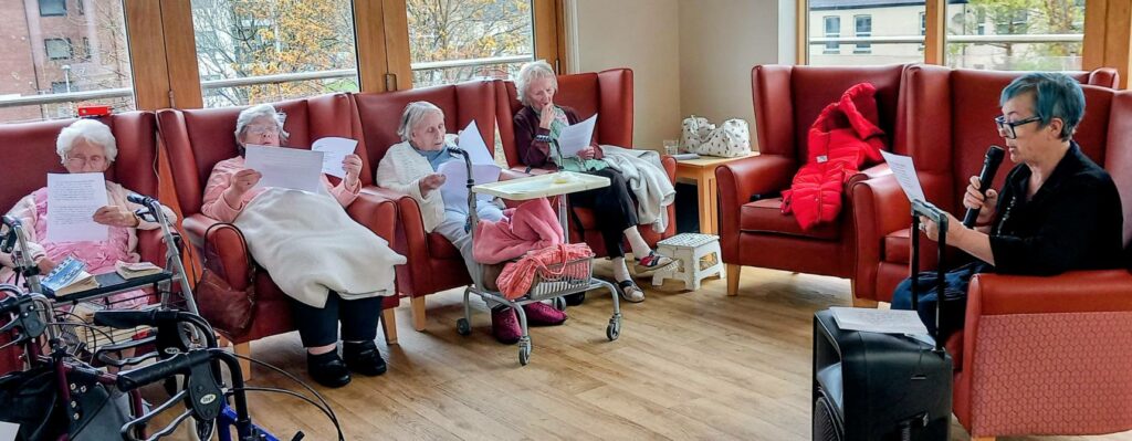A group of elderly women sit in armchairs, holding papers, whilst another woman uses a microphone to read or sing to them. They share meaningful time together in a bright communal room.