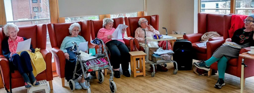 Five elderly women sit in armchairs in a bright room, sharing a reflective moment together as they hold papers and enjoy uplifting activities. Mobility aids and personal belongings are close by.