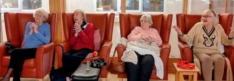 Four elderly women are seated in armchairs, some with percussion instruments on their laps, enjoying feel-good fun and a thoughtful time together in a bright room.