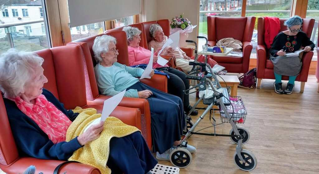 Five elderly women enjoy Thoughtful Time in armchairs, holding papers; one has a walking frame. Another woman sits by the window, also reading. The bright room, with large windows and wooden floors, reflects their time together.