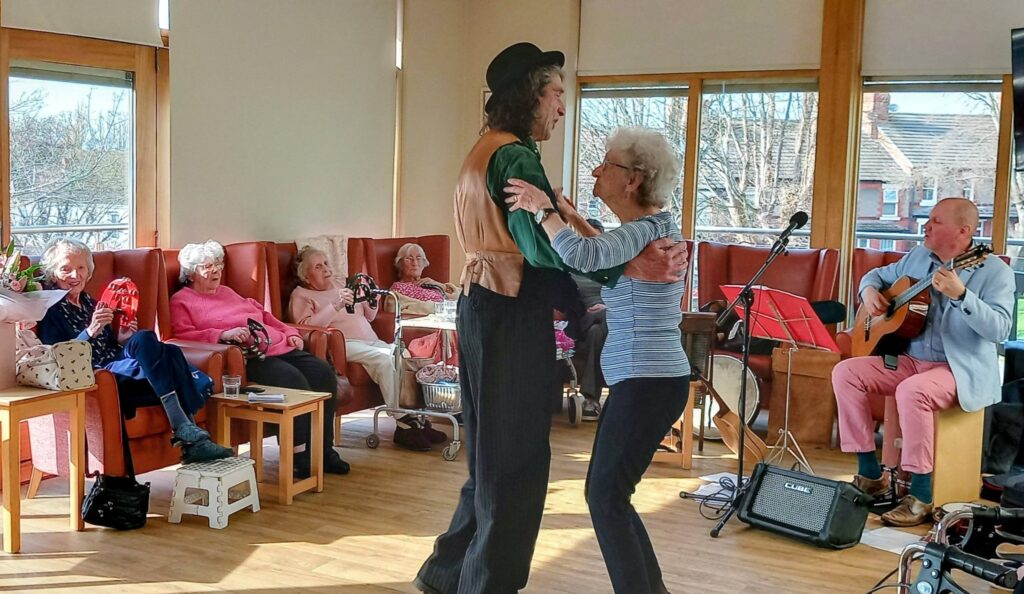 Elderly people sit smiling as a man and woman dance in time, enjoying live guitar music in a bright room.