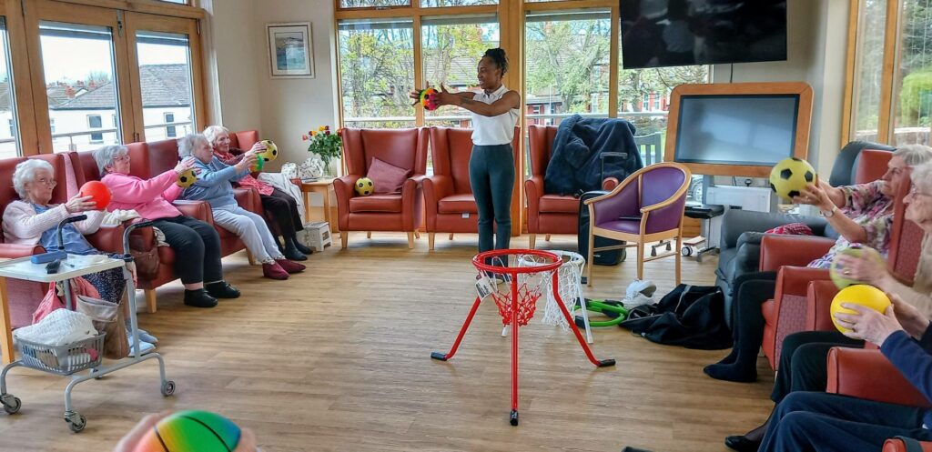 A group of elderly people sit in a circle in a bright room, taking part in a ball toss activity with a facilitator and a basketball hoop in the centre.