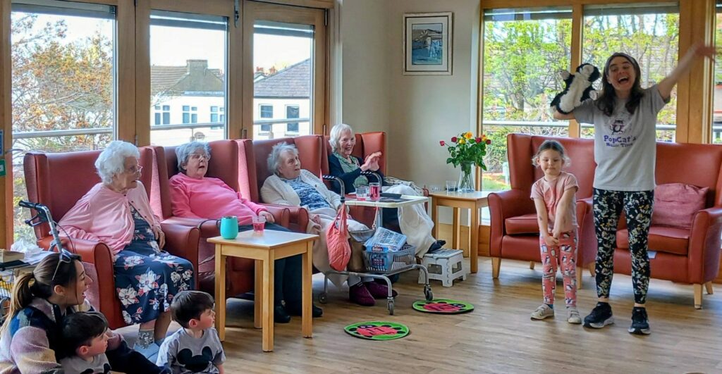 A group of elderly women and children observe a woman holding a small animal as she stands with a young girl in a bright room with large windows.