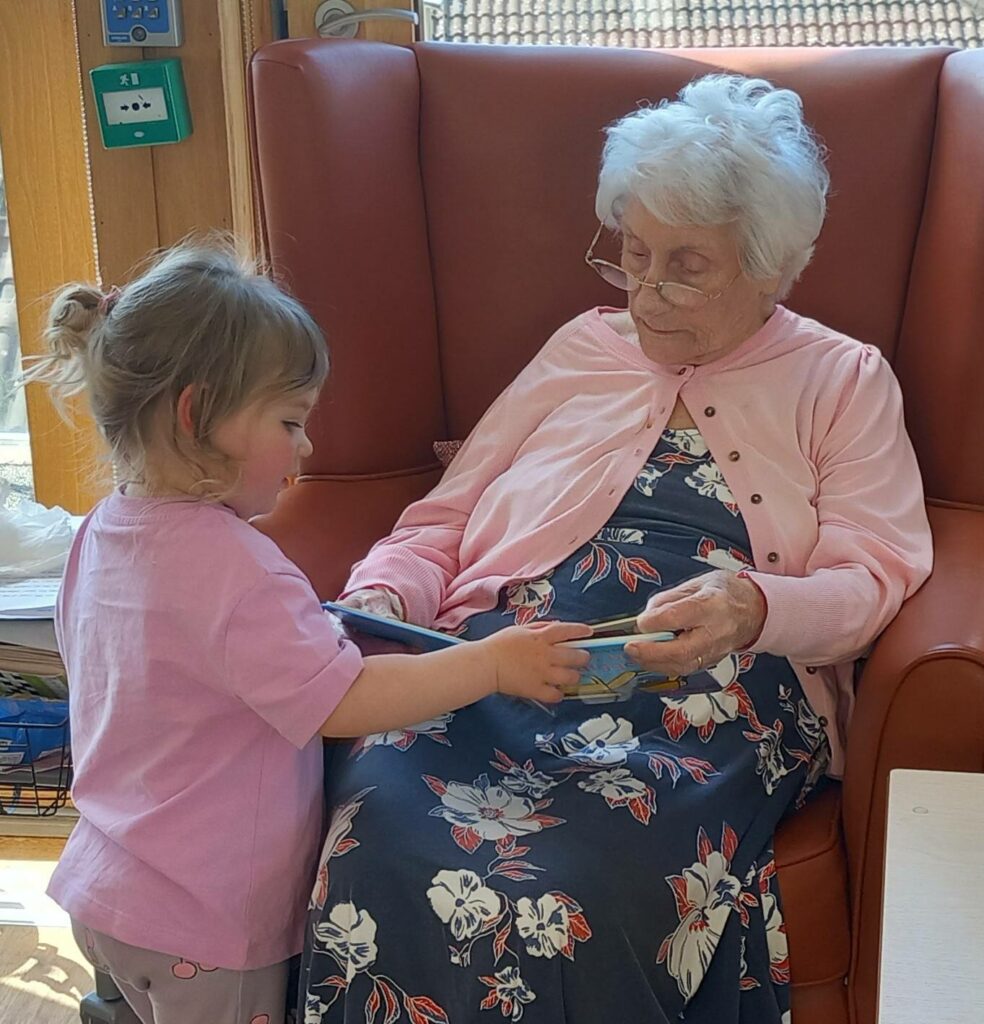 A young girl hands a book to an elderly woman seated in a chair; both are wearing pink tops.