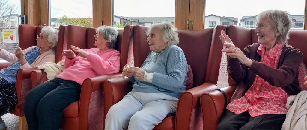 Four elderly women sit in a row of armchairs, smiling and raising their hands as if taking part in an activity, inside a bright room with large windows.