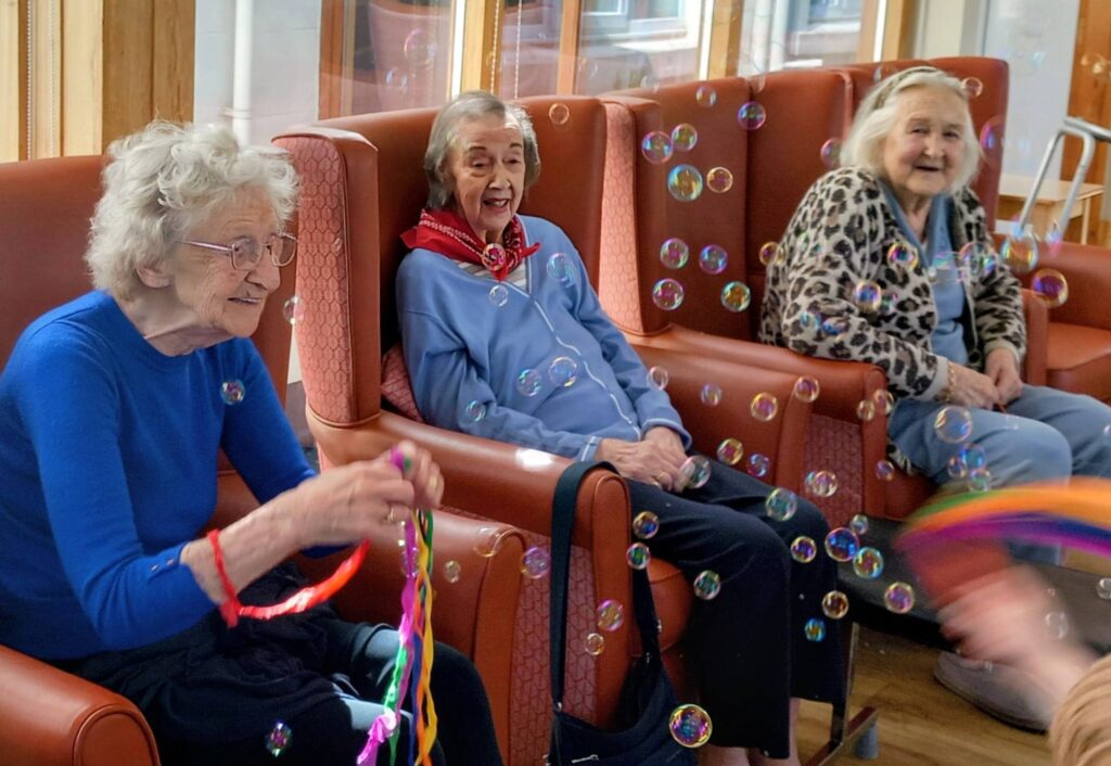 Three elderly women sit in armchairs indoors, smiling as they watch bubbles drifting about. One woman holds colourful ribbon streamers.