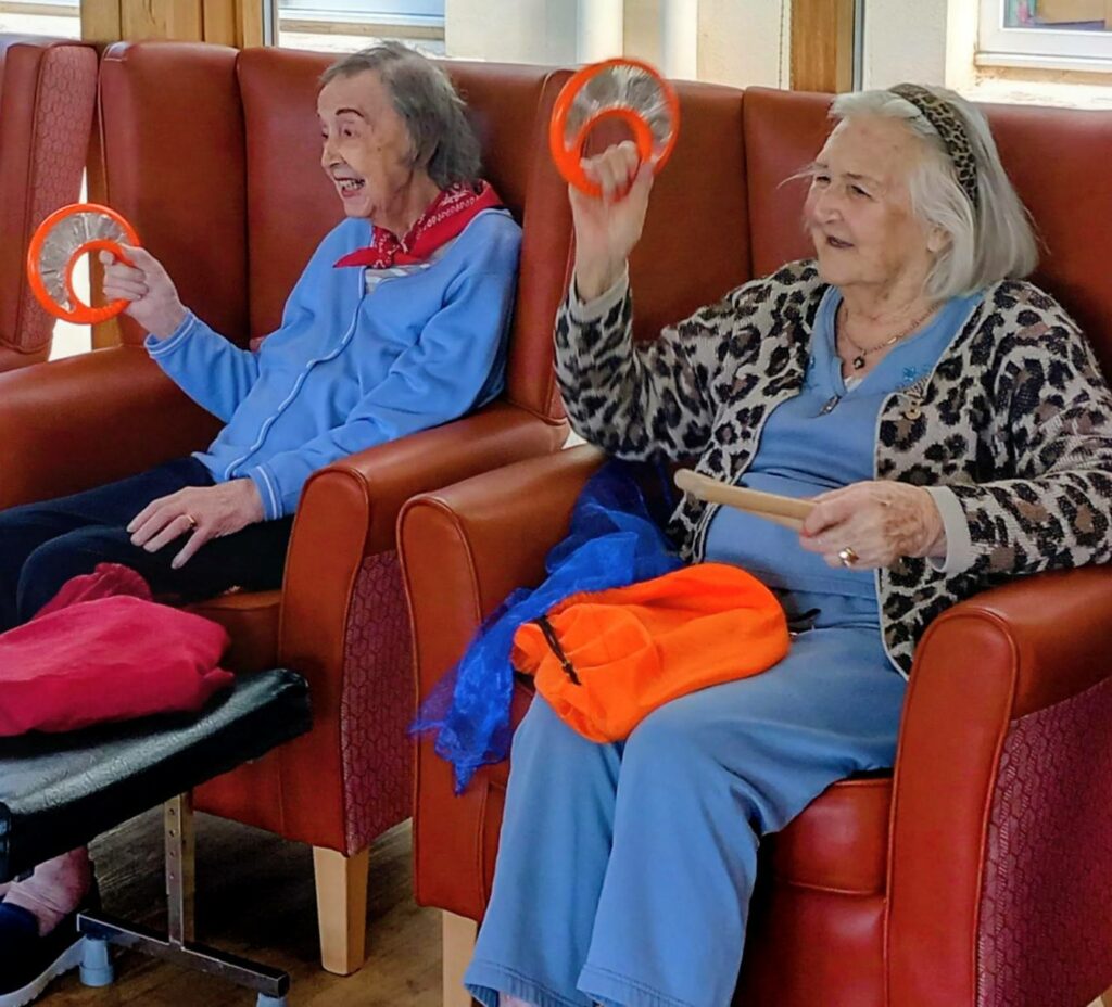 Two elderly women sit in armchairs, smiling and holding orange hand drums, taking part in an indoor group activity.