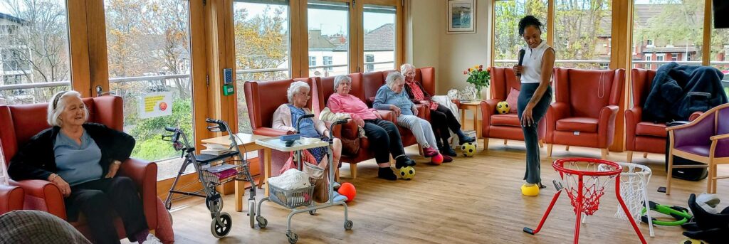 A group of elderly people sit in armchairs, watching a woman standing and playing an indoor basketball game in a bright communal lounge.